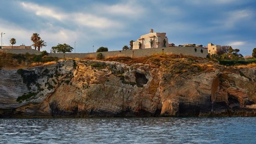 Building on a cliff under a cloudy sky next to the sea, boat trip around the island of Ortigia,