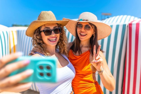 Two cheerful women wearing stylish sunglasses and trendy sun hats are capturing a joyful selfie on