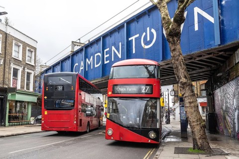 Red double decker buses driving on a london street, passing under a bridge with the camden town