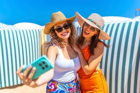 Two cheerful women wearing summer hats and sunglasses are taking a selfie on a sunny beach with