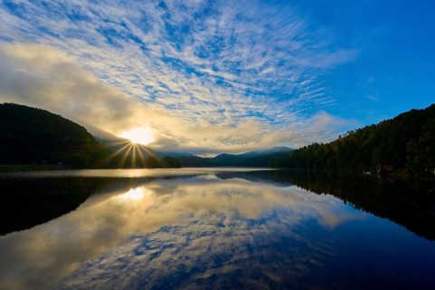 The early morning shows sunbeams through clouds reflecting on a calm lake in a mountainous