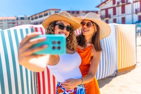 Two smiling women with sun hats and sunglasses taking a selfie on a sunny beach with colorful