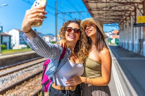 Two happy female tourists taking a selfie with smartphone at a train station, enjoying their summer