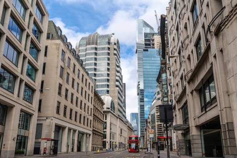 Red double decker bus driving on empty street surrounded by modern and historic buildings in city