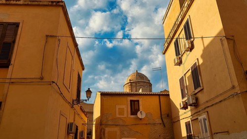 Narrowed view of a dome, framed by yellow houses and cables, with partly cloudy sky, Ortigia