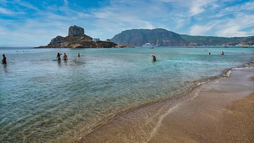 Clear sea with people bathing, surrounded by an island and mountains under a cloudy sky, Agios