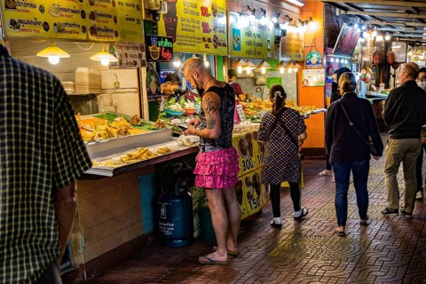 Tatooed man in pink skirt and flip flops ordering food from a street vendor in the Night Market in