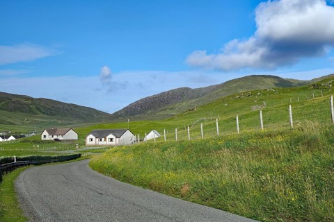 Winding country road leads through a rural area with meadows and houses under a blue sky, Barra,