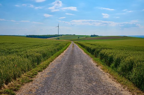 A gravel path leads through green fields with a wind turbine on the horizon under a blue sky,
