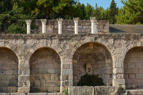 Ancient ruins with arches and columns, surrounded by forest and vegetation, retaining wall with