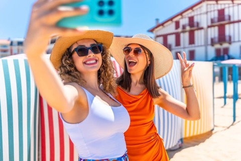 Two cheerful women wearing sun hats and sunglasses, capturing a fun selfie on a sunny beach,