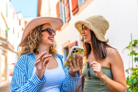 Two happy women wearing summer hats and sunglasses are using a smartphone and laughing while
