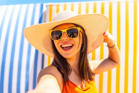 Young woman with sunhat and sunglasses taking a selfie on a striped background at the beach during