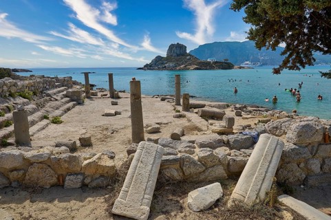 Ancient ruins in front of a rocky coast with a view of the clear sea and a small, mountainous