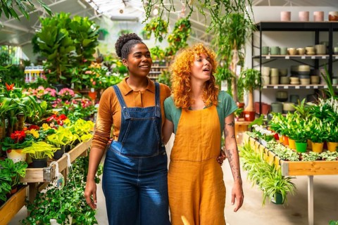 Two young women walking arm in arm through a garden center, discussing their work among vibrant