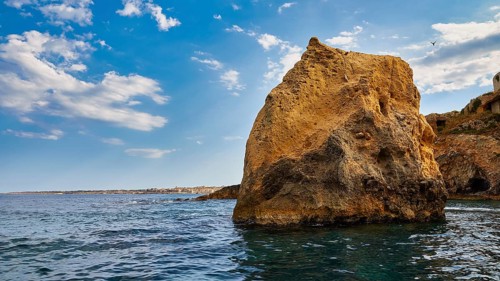 A large rock in the sea under a blue sky with white clouds, boat trip around the island of Ortigia,