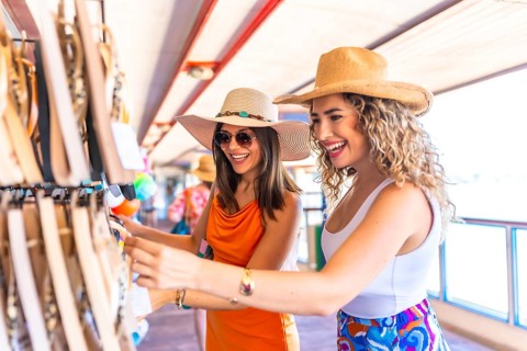Two smiling women wearing summer hats and sunglasses are choosing souvenirs at a seaside shop while