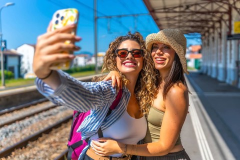 Two female tourists with backpack and straw hat are taking a selfie with smartphone at a train