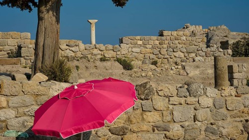 Ancient ruins with a red parasol in the foreground under a clear blue sky, Agios Stefanos Beach,