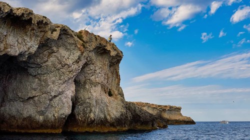 Person fishing from a rocky cliff under a cloudy sky, boat trip around the island of Ortigia, Isola