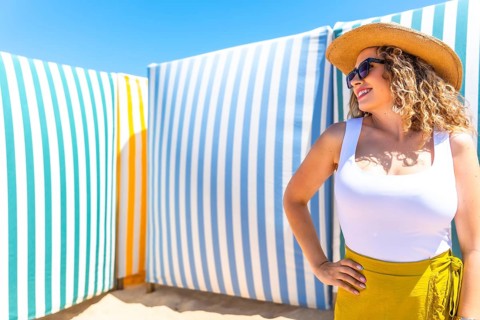 Young woman with curly hair, sunglasses and straw hat posing on beach with colorful striped cabanas