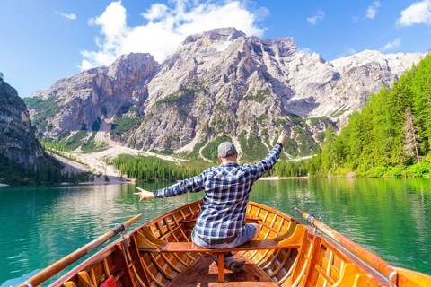 A man sits in a wooden boat and spreads out his arms as he heads towards an impressive mountain,