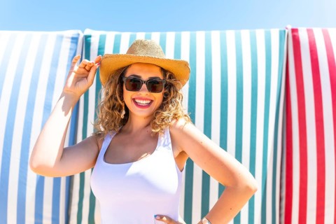 Cheerful young woman with curly hair, wearing sunglasses and a straw hat, smiles while posing