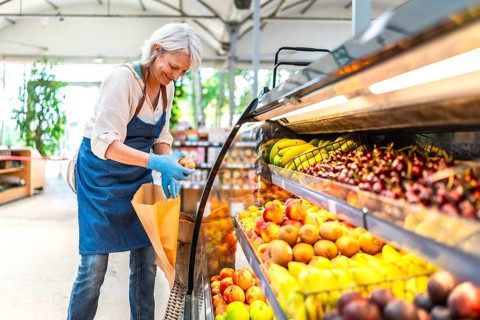 Smiling senior saleswoman wearing gloves, arranging vibrant fresh fruits in a supermarket display