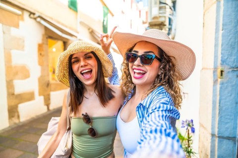 Two cheerful female tourists wearing summer hats and sunglasses are taking a selfie with a cheerful