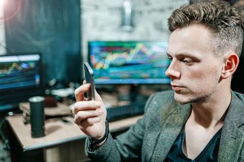 Man focused on a smartphone in an office with trading charts on monitors