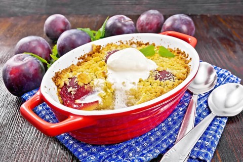 Crumble with plum and ice cream in a red brazier with spoons on a napkin, plums and mint on dark