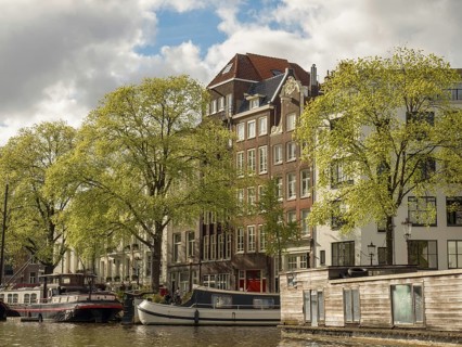 Building fronts along a canal with boats in the water, surrounded by green trees and a cloudy sky,