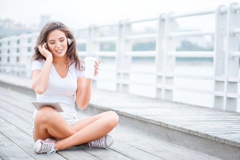 Happy young woman with music headphones and a cup of coffee to go, surfing the internet on a tablet