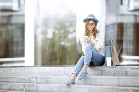 Cheerful young woman with a disposable coffee cup and shopping bags, sitting on the stairs and
