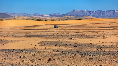 An off-road vehicle moves through a vast landscape of sand dunes and mountains, With off-road