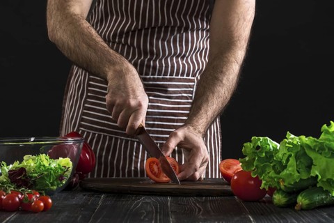 Man in a striped apron preparing salad on a wooden table. Men's hands cut the tomato to make a