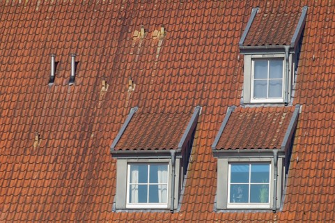 Close-up view of a sloped roof featuring red clay tiles, three dormer windows, and two chimneys