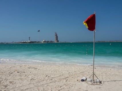 A red warning sign stands on the beach with turquoise water and a blue sky in the background,