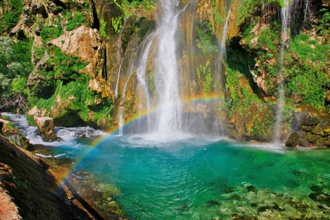 Waterfall with rainbow, surrounded by rocks and green foliage under a clear sky, Topoljski Buk