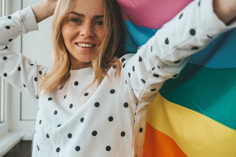 Young blonde millennial hippie woman with rainbow LGBTQ flag at home balcony. Peace and freedom the