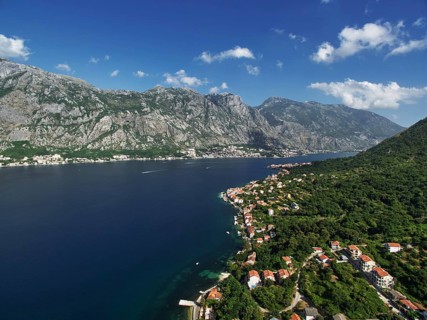 Resort town on the shores of the Bay of Kotor among green trees at the foot of the mountains.