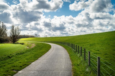 An empty country lane winds along green meadows and a fence under a cloudy sky, cycle path, Leer,