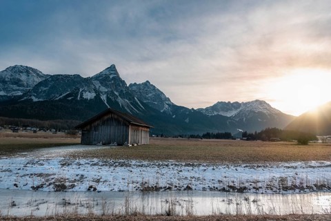 Winter alpine landscape with a wooden cabin, a frozen river and the snowy Alps mountains at sunset,