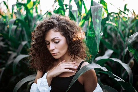 A serene woman with curly hair touches her chest in a sunlit cornfield