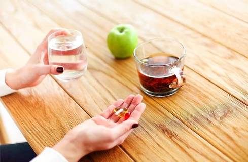 High angle of anonymous female with pills and cup of water witting at table with tea and apple
