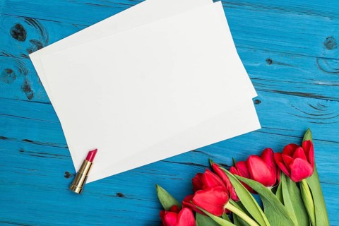 Red tulips, lipstick and a white sheet of paper for your greetings on the background of blue wooden