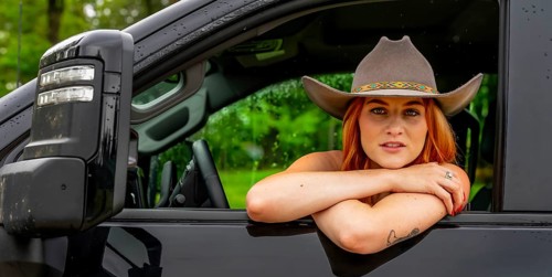 A beautiful redhead model sits in her truck while wearing a cowboy hat on her farm