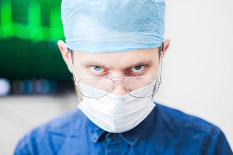 A surgeon with an intense expression, wearing mask and glasses, in a blurred clinical background