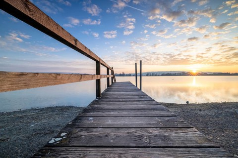 A wooden footbridge leading onto a calm lake as the sun sets under a cloudy sky, Naturfreundehaus,