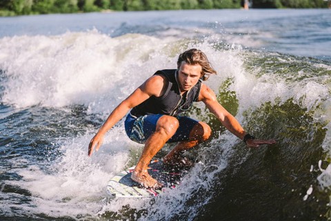 Beautiful long haired blond wakesurf in vest on board along waves of lake. Athletic male athlete